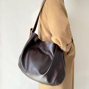 May include: A dark brown leather shoulder bag with a long strap. The bag has a slouchy, hobo-style design and a smooth texture. The strap is a darker brown and appears to be adjustable. The bag is worn over the shoulder.
