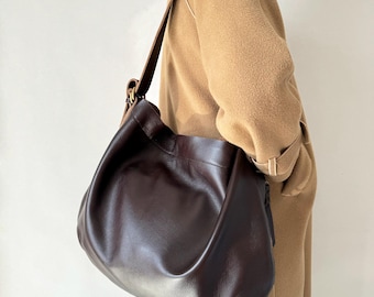 May include: A dark brown leather shoulder bag with a long strap. The bag has a slouchy, hobo-style design and a smooth texture. The strap is a darker brown and appears to be adjustable. The bag is worn over the shoulder.