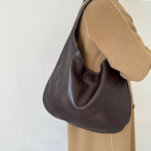 May include: A dark brown leather shoulder bag with a single strap. The bag has a rounded shape and a button closure. The bag is being worn over a tan coat.