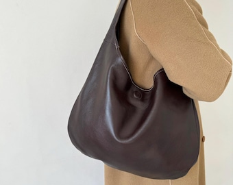 May include: A dark brown leather shoulder bag with a single strap. The bag has a rounded shape and a button closure. The bag is being worn over a tan coat.