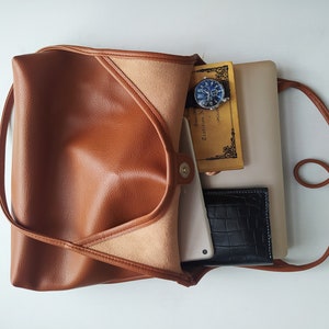 Puede incluir: Un bolso tote de cuero marr&oacute;n con cierre de solapa y una correa larga. El bolso est&aacute; abierto y contiene un port&aacute;til, un cuaderno, un reloj y una cartera.