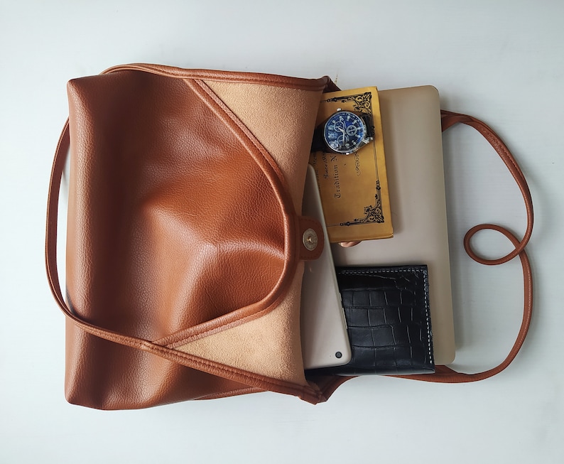 Puede incluir: Un bolso tote de cuero marr&oacute;n con cierre de solapa y una correa larga. El bolso est&aacute; abierto y contiene un port&aacute;til, un cuaderno, un reloj y una cartera.