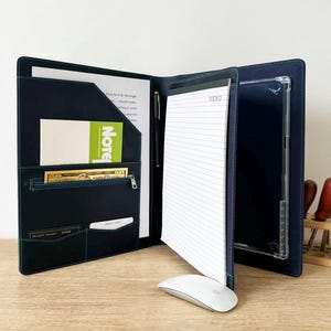 May include: A dark blue leather portfolio organizer featuring a notepad with the word MEMO, a pen holder, a zippered pocket, card slots, and a clear sleeve for a tablet. The interior includes a green note card and a fifty dollar bill insert.