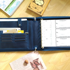 May include: A blue leather binder with a zipper closure, a pocket for a tablet, and a section for pens and cards. The binder has a ring mechanism and is open to reveal blank cheque forms.