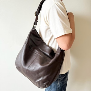 May include: A dark brown leather shoulder bag with a textured surface. The bag has a wide, adjustable strap and a large, slouchy design. The bag is worn over the shoulder.
