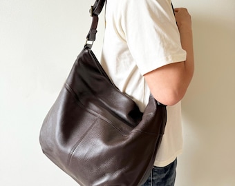 May include: A dark brown leather shoulder bag with a textured surface. The bag has a wide, adjustable strap and a large, slouchy design. The bag is worn over the shoulder.