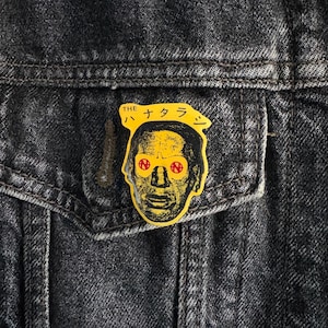 Könnte beinhalten: Eine gelbe und schwarze Anstecknadel mit einem Comic-Gesicht und dem Text "THE ハナタラジ" in Japanisch.