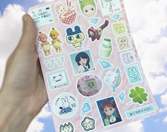 May include: A pink sheet of paper with various stickers on it. The stickers feature cartoon characters, anime characters, and Japanese text. Some of the text includes "OTL 좌절금지 ㅠㅂㅠㅠ 우울금지 이재" and "꽃이 예쁘게 피었다".