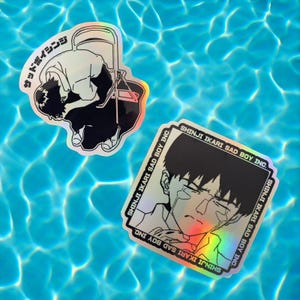 Pode incluir: Dois adesivos holográficos com um personagem do anime Neon Genesis Evangelion. O primeiro adesivo mostra o personagem Shinji Ikari sentado em uma cadeira com o texto "Sad Boy Inc." e "Shinji Ikari Sad Boy Inc." O segundo adesivo mostra um close-up do rosto de Shinji Ikari com o texto "Shinji Ikari Sad Boy Inc." repetido quatro vezes.
