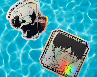 May include: Two holographic stickers featuring a character from the anime Neon Genesis Evangelion. The first sticker shows the character Shinji Ikari sitting on a chair with the text "Sad Boy Inc." and "Shinji Ikari Sad Boy Inc." The second sticker shows a close-up of Shinji Ikari's face with the text "Shinji Ikari Sad Boy Inc." repeated four times.