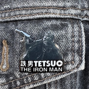 Peut inclure: Épinglette métallique noire et argentée représentant une figurine et le texte "TETSUO THE IRON MAN" en blanc. L'épinglette est fixée à une veste en jean. Il s'agit d'un objet de collection.