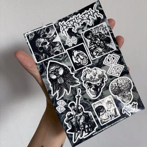 May include: A sheet of black and white stickers featuring various illustrations from the manga series Berserk. The stickers include characters, creatures, and symbols from the series. The sheet is printed with a black and white marbled background.