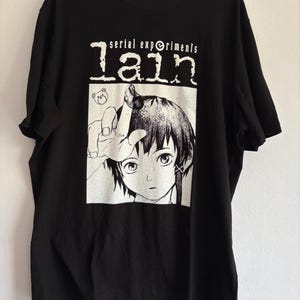Può includere: T-shirt nera con grafica bianca di un personaggio anime e la scritta "serial experiments lain". L'illustrazione mostra una persona con i capelli corti che fa un gesto con la mano.