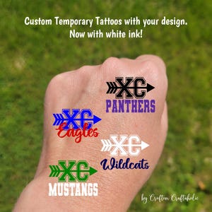 May include: Four temporary tattoos applied to skin, each featuring an arrow design with the letters XC and a team name. The designs include black Panthers, blue Eagles, white Wildcats, and green Mustangs, with text above reading Custom Temporary Tattoos with your design. Now with white ink!