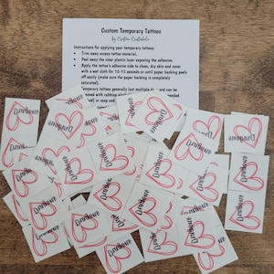 May include: A pile of white temporary tattoos with a pink heart and the word "Dradove" printed on each. The tattoos are arranged in a random pattern. The tattoos are on a wooden surface.