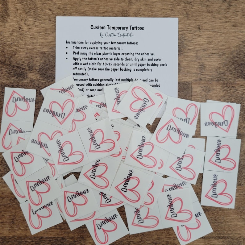 May include: A pile of white temporary tattoos with a pink heart and the word "Dradove" printed on each. The tattoos are arranged in a random pattern. The tattoos are on a wooden surface.