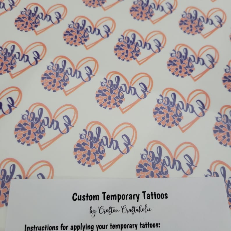 May include: A sheet of temporary tattoos with a heart design and the word "Eagles" in purple script. The heart is outlined in orange and filled with a purple and orange pom-pom. The text "Custom Temporary Tattoos by Crofton Craftaholic" is at the bottom of the sheet. Instructions for applying the tattoos are also included.