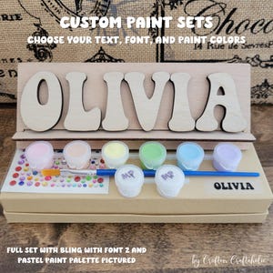 May include: A wooden sign with the name "OLIVIA" painted in a whimsical font. The sign is part of a craft kit that includes a set of pastel paint pots, a paintbrush, and a bag of colorful rhinestones.