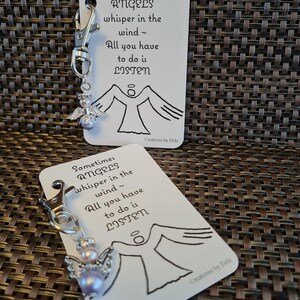 Op de afbeelding: Een witte kaart met de tekst "Sometimes Angels whisper in the wind - All you have to do is LISTEN" en een zwart-witte tekening van een engel met vleugels. De kaart is bevestigd aan een zilveren sleutelhanger met een kleine roze engelbedel.