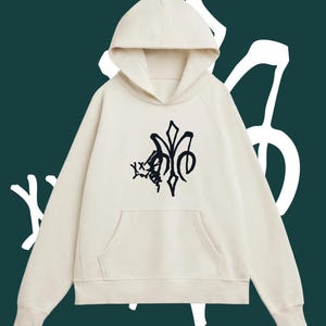 May include: Cream-colored hoodie with a black graphic design on the front. The hoodie features a hood, long sleeves, and a front pocket. The background is a dark teal color with white abstract shapes.