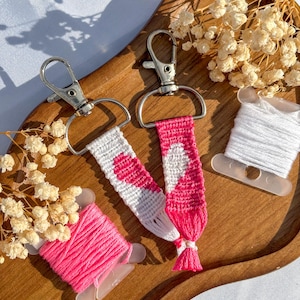 Peut inclure: Deux porte-clés faits à la main avec des fermoirs argentés, présentant un motif de cœur blanc et rose. Les porte-clés sont présentés sur un plateau en bois avec des bobines de fil rose et blanc et des fleurs séchées. Les porte-clés ont un pompon à l'extrémité.