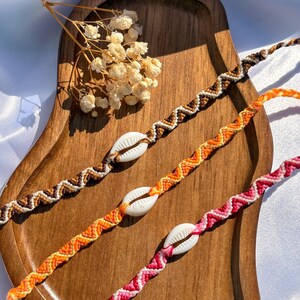 Peut inclure: Trois bracelets d'amitié faits à la main avec des accents de coquillages. Les bracelets présentent des motifs en chevrons marron, orange et rose, présentés sur un plateau en bois. Une brindille de fleurs séchées ajoute une touche naturelle.