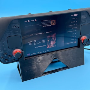 Könnte beinhalten: Ein schwarzes Handheld-Gaming-Gerät mit roten Knöpfen und einem Bildschirm, auf dem ein Spiel namens "Deathsmiles I" mit dem Text "MY ADVENTURES" und "Fortress? Beast?" angezeigt wird. Das Gerät steht auf einem schwarzen Ständer.