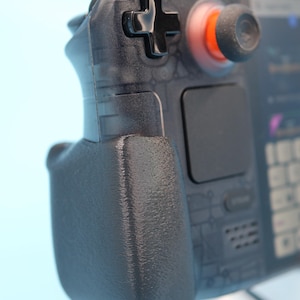 May include: Close-up of a handheld gaming device with a black D-pad and thumbstick. The thumbstick has an orange ring around it. The device has a textured grip and a screen visible in the background. The device is black and gray.