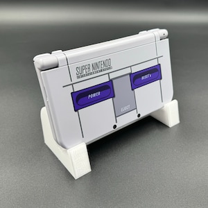 May include: A white Nintendo Switch console with a custom Super Nintendo Entertainment System design. The console has a white stand and features buttons labeled "Power", "Eject", and "Reset".
