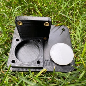 May include: A black 3D printed holder with a round opening and a white disc. The holder has two gold screws on the side.