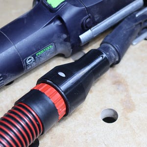 Könnte beinhalten: Ein schwarzes und silbernes Elektrowerkzeug mit einem schwarzen Schlauch und roten Akzenten. Das Werkzeug hat eine grüne Taste und die Marke "Festool" ist sichtbar. Der Schlauch ist mit dem Werkzeug verbunden und hat ein geripptes Design.