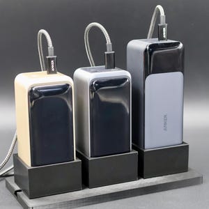 May include: Three black portable power banks with different colored casings: beige, gray, and dark gray. Each power bank has a black charging cable attached and is sitting on a black stand. The power banks have the word "ANKER" printed on them.