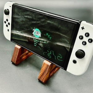 Może przedstawiać: Biało-czarna konsola Nintendo Switch OLED wyświetlająca menu gry Zelda, spoczywająca na drewnianym stojaku. Ekran pokazuje tytuł gry i opcje menu w kolorze zielonym i białym. Stojak wykonany jest z ciemnobrązowego drewna.