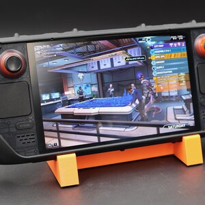 May include: A black handheld gaming device with a screen displaying a video game. The device has two red joysticks and a black button on the left side. The screen shows a game with a blue pool table and characters in a futuristic setting. The text "SATURDAY" is visible on the screen.