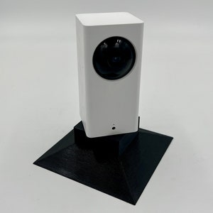 Op de afbeelding: Een witte beveiligingscamera met een zwarte lens staat op een zwarte, driehoekige basis.