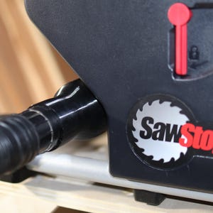 May include: Close-up of a black SawStop table saw with a dust port and a red lever. The SawStop logo is visible on the side of the saw.