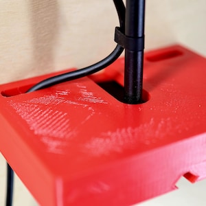 May include: A bright red, rectangular, 3D-printed object with a hole and a slot for a black cable and a black pole. The object is mounted on a light-colored wooden surface. The cable is secured with a black zip tie.