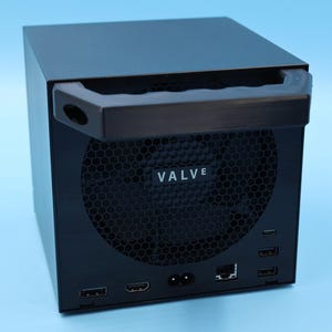 May include: A black, cube-shaped computer console featuring a prominent top handle and a large circular honeycomb ventilation grille on the front. The word VALVE is printed in white on the centre of the grille, with various ports visible below.
