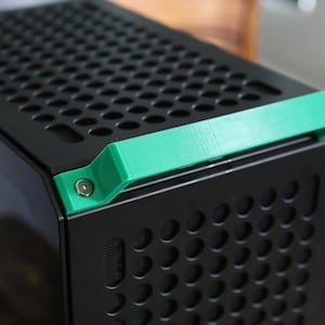 Könnte beinhalten: Ein schwarzes Computergehäuse mit perforierter Oberseite und Seitenwänden. Ein hellgrüner Griff ist mit einer silbernen Schraube an der Oberseite des Gehäuses befestigt. Das Gehäuse hat ein modernes, minimalistisches Design.