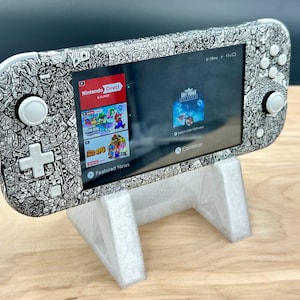 May include: A white Nintendo Switch Lite console with a black and white doodle design is displayed on a white plastic stand. The console screen shows the Nintendo Direct logo and a game called "Sky: Children of the Light".