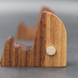 Peut inclure: Gros plan sur un support de téléphone en bois clair, présentant un design incurvé et un grain naturel. Il est orné d'une petite cheville circulaire. Le fond est gris uni.