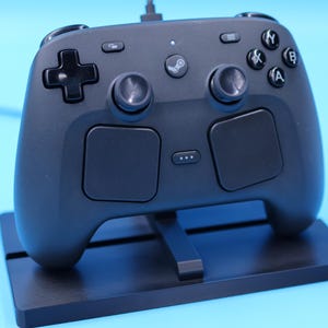 May include: A matte black video game controller featuring two analog sticks, a directional pad, four action buttons labeled A, B, X, and Y, two large touchpads, and a central Steam logo button, resting on a black rectangular display stand.