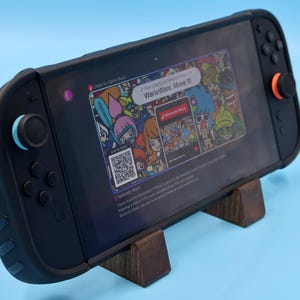 May include: A black handheld gaming console with a screen displaying the WarioWare: Move It! game interface, featuring a QR code and colorful character graphics. The console has a black protective case, one orange joystick, and one light blue joystick.