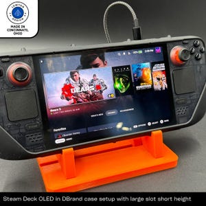 Peut inclure: Une console de jeu Steam Deck OLED noire et orange avec un étui DBrand et un support. La console affiche le jeu Gears 5 sur l'écran. Le support est orange et possède une large fente pour la console.