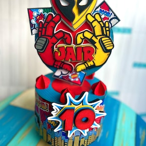 May include: A red and yellow cake topper with a heart-shaped design featuring a superhero mask. The topper says "JAIR" and has a comic book style. The cake is decorated with a red number 10 and a blue and yellow comic book design.