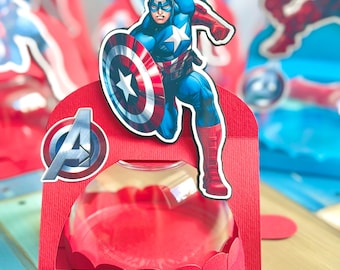 Könnte beinhalten: Eine rote Geschenkbox aus Papier mit einem Ausschnitt von Captain America, der einen Schild hält, einem transparenten Kunststoffkuppeleinsatz und einem Avengers-Logo-Aufkleber auf der linken Seite. Die Box hat eine strukturierte Oberfläche.