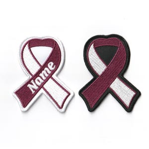 May include: Two embroidered iron-on patches in the shape of a burgundy and white ribbon. One patch has a white background and the other has a black background. The burgundy ribbon has the word "Name" embroidered on it.