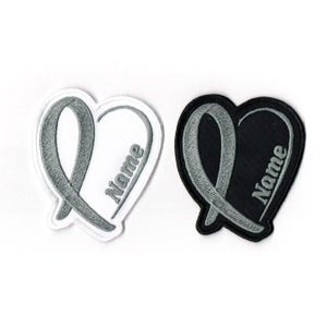 May include: Two heart-shaped patches. One is white with a gray ribbon design and the word "Name" embroidered in gray. The other is black with a gray ribbon design and the word "Name" embroidered in gray. Patches are likely for clothing or accessories.