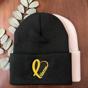 May include: Black beanie with a gold embroidered heart and the word "Name". A second, cream-colored beanie is visible behind the black one. The beanies are displayed on a dark wood surface with green leaves.