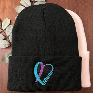 May include: Black beanie with a teal and purple ribbon heart design and the name "Name" embroidered in teal. A second, off-white beanie is partially visible behind the black one. The beanies are made of a soft, knitted material.